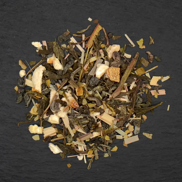 Un amestec de ceai verde, bucăți de fructe uscate, lemongrass și alte plante, pe o suprafață închisă la culoare.