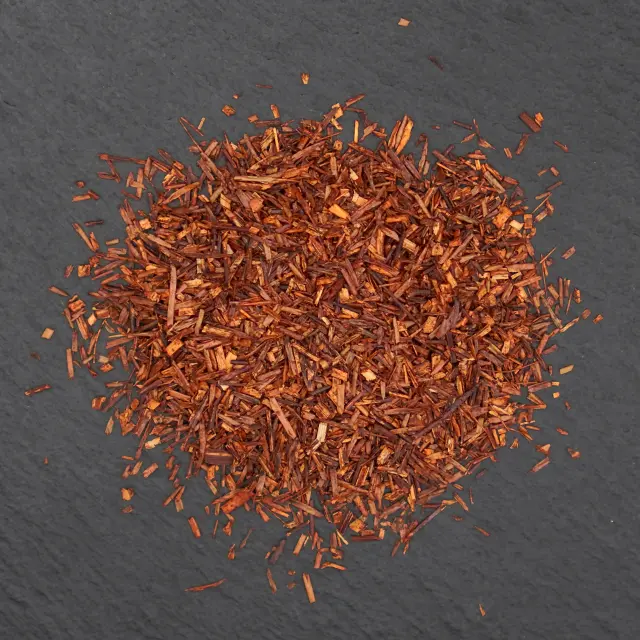 O grămadă de ceai rooibos original, uscat, de culoare roșu-maroniu, pe o suprafață închisă la culoare.
